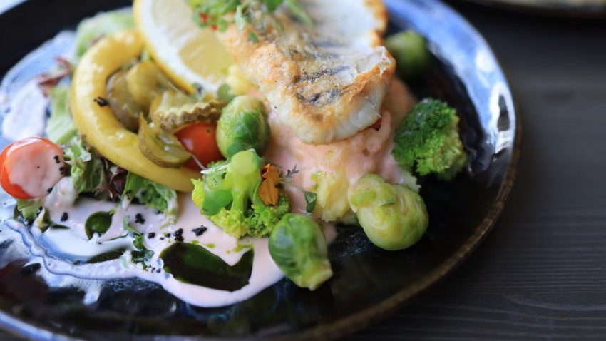 Close-up of a gourmet fish dish with assorted vegetables on a dark plate.